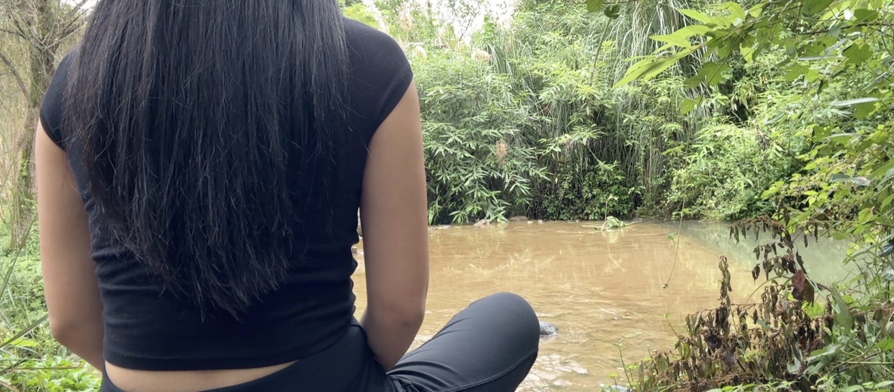 Dami sitting quietly by a river, seen from behind
